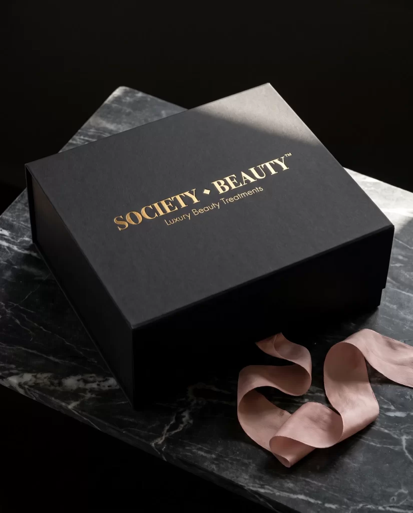 Zwarte geschenkdoos van Society Beauty met logo in goudfolie en een roze lint op marmer